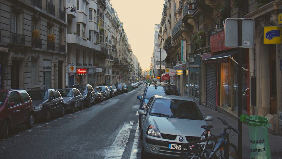 A narrow Paris street in pale evening light, cars parked along both sides.