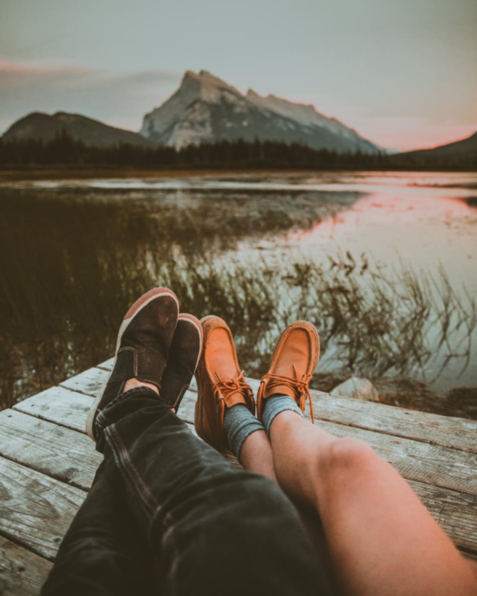 Two pairs of feet on a wooden dock at dusk, a mountain and lake behind in warm, faded colour.